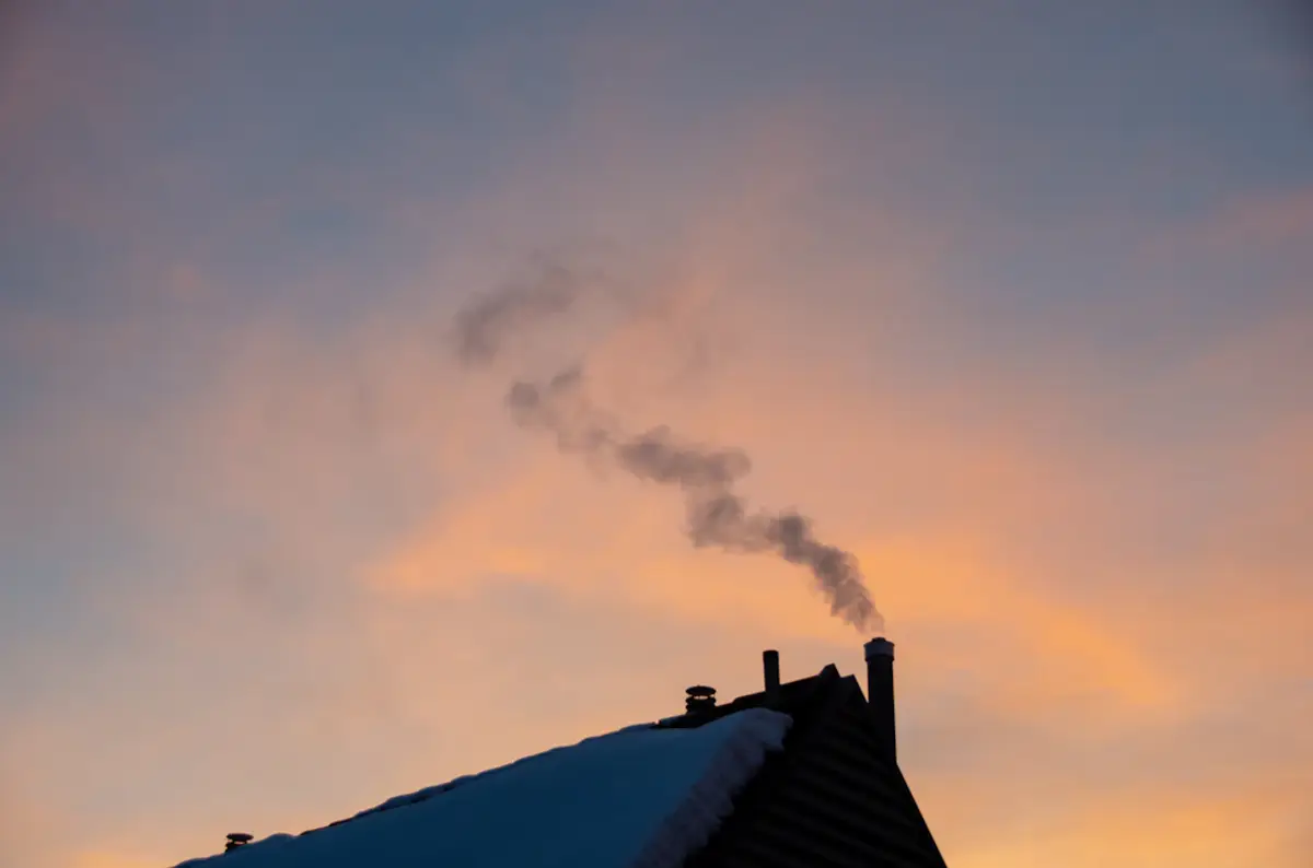smoke rising from a chimney on a snow-covered roof during sunset, indicating home heating in winter